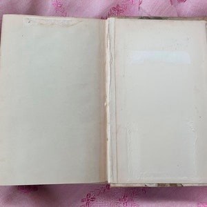 May include: An open book with aged, off-white pages. The book's spine is visible, and the edges of the pages show signs of wear. A white label is affixed to the right page. The book rests on a pink floral patterned surface.