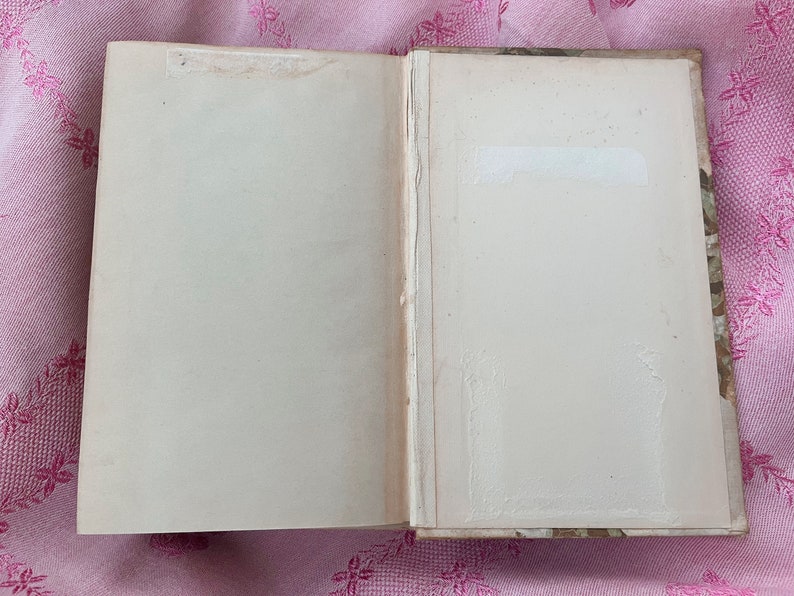 May include: An open book with aged, off-white pages. The book's spine is visible, and the edges of the pages show signs of wear. A white label is affixed to the right page. The book rests on a pink floral patterned surface.