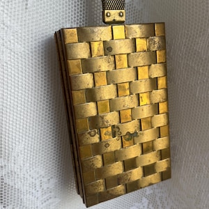 May include: A vintage gold-toned metal compact with a woven basket weave design. The rectangular compact has a matching chain handle. The compact is slightly tarnished, showing its age. The clasp is visible on the front.