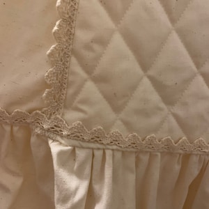 Puede incluir: Primer plano de una tela acolchada color crema con un patr&oacute;n de diamantes. El borde est&aacute; adornado con un delicado encaje festoneado. Debajo de la secci&oacute;n acolchada, una tela fruncida a&ntilde;ade un elemento suave y texturizado.