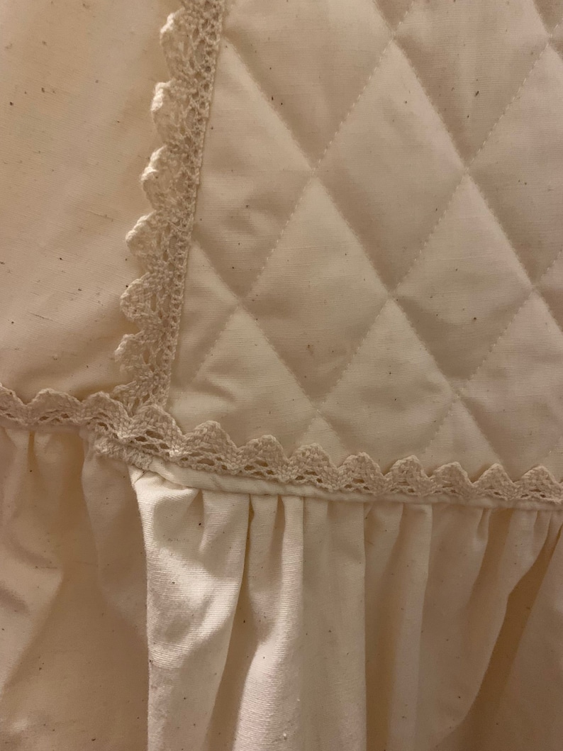 Puede incluir: Primer plano de una tela acolchada color crema con un patr&oacute;n de diamantes. El borde est&aacute; adornado con un delicado encaje festoneado. Debajo de la secci&oacute;n acolchada, una tela fruncida a&ntilde;ade un elemento suave y texturizado.