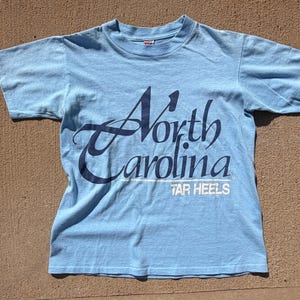 以下が含まれることがあります： 胸元に濃い青色の筆記体でNorth Carolinaと書かれ、その下に白いブロック体でTAR HEELSとプリントされた、水色の半袖Tシャツ。シャツの前面には、スタイリッシュなスクリプトフォントでテキストが配置されています。
