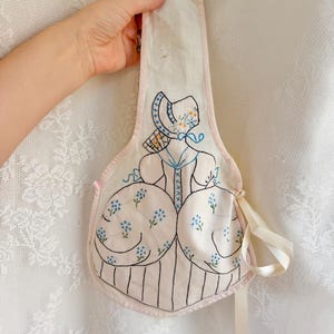 May include: A white fabric pouch with a long handle, featuring an embroidered design of a figure in a bonnet and wide skirt. The embroidery uses black, blue, and yellow thread to depict the figure, accented with small blue floral motifs and a cream ribbon.