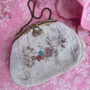 Puede incluir: Bolso de mano vintage de cuentas color crema con un diseño floral en tonos rosa, azul y marrón. El bolso tiene un marco y un asa dorados. El bolso se encuentra sobre un fondo de tela rosa floral.