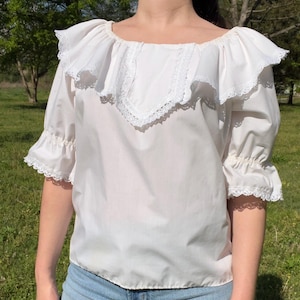 May include: A white blouse with a ruffled neckline and sleeves. The blouse features a square neckline with a ruffled collar and lace trim. The sleeves are three-quarter length with ruffled cuffs and lace trim. The blouse is made of a lightweight fabric.