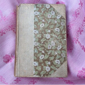May include: A vintage book with a floral design on the cover. The cover features a pattern of white flowers and green leaves on a brown background. The spine and corners have an ornate, cream-colored design.