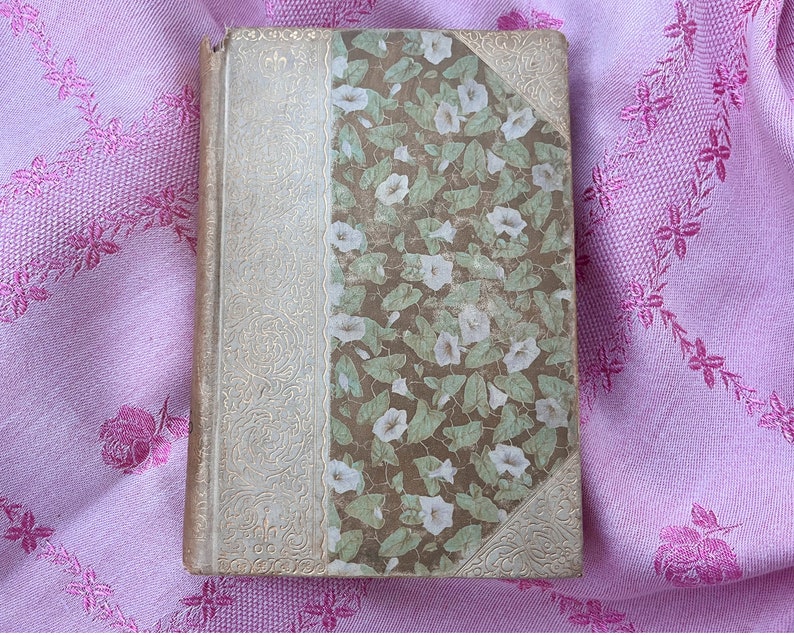May include: A vintage book with a floral design on the cover. The cover features a pattern of white flowers and green leaves on a brown background. The spine and corners have an ornate, cream-colored design.