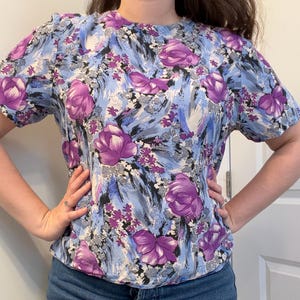 May include: A short-sleeved blouse with a floral design. The top has a light blue backdrop with large purple flowers and smaller purple and grey floral accents. The blouse has a round neckline.