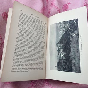 May include: An open book with text and a black and white image of a city on a hillside. The book is open to page 20, with the title "THE ALHAMBRA" visible. The book is on a pink floral patterned fabric.