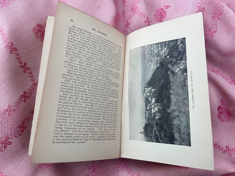 May include: An open book with text and a black and white image of a city on a hillside. The book is open to page 20, with the title "THE ALHAMBRA" visible. The book is on a pink floral patterned fabric.