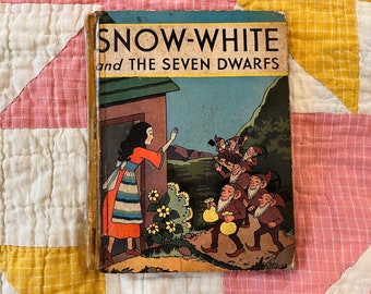 May include: Vintage hardcover book titled "Snow-White and the Seven Dwarfs." The cover features an illustration of Snow White waving from a doorway, with the seven dwarfs approaching. The book has a worn appearance.