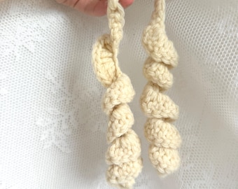 May include: A cream-colored crochet accessory featuring two long, spiraled, corkscrew-shaped yarn strands connected by a single looped top section. The item is crafted from thick, textured yarn with a consistent off-white hue throughout the design.