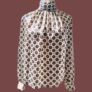 May include: A long-sleeved blouse featuring a high mock neck collar and a repeating geometric pattern in dark red and black on a cream-colored background. The garment has gathered fabric at the neckline and buttoned cuffs on the long sleeves.
