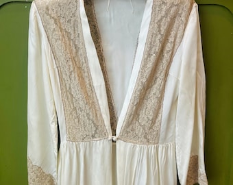 May include: Ivory-colored vintage robe with lace detailing. The robe features a V-neck design with lace panels on the front and sleeves. It hangs on a white hanger against a green background. The robe has a button closure at the waist.