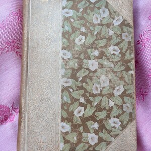 May include: An antique book with a floral design. The spine and corners are a light cream color with gold filigree. The cover features a brown background with a pattern of white flowers and green leaves. The book is closed and appears to be in good condition.