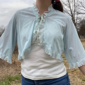May include: A light blue, sheer bolero with ruffled edges and three-quarter sleeves. The bolero has a delicate tie closure at the neckline and is worn over a white top. The garment is made of a lightweight fabric.