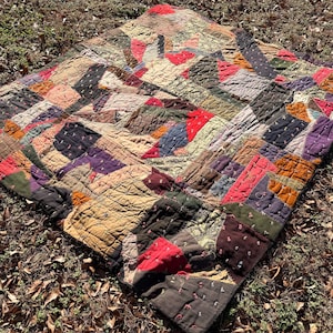 May include: A patchwork quilt with a variety of colors including red, purple, green, and tan. The quilt is made of many small, irregularly shaped pieces of fabric sewn together. The quilt is lying on the ground.