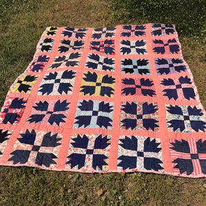 May include: A handmade quilt with a repeating pattern of navy blue and patterned blocks set against a coral background. The quilt is spread out on a grassy surface, showcasing its intricate design and craftsmanship. The quilt's edges are finished with a floral patterned fabric.