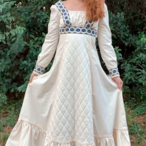 Puede incluir: Vestido largo color crema con mangas largas, cuello cuadrado y corpi&ntilde;o acolchado. El vestido presenta detalles bordados en azul y blanco en el escote, la cintura y los pu&ntilde;os. La falda tiene un dobladillo con volantes, creando una silueta fluida.