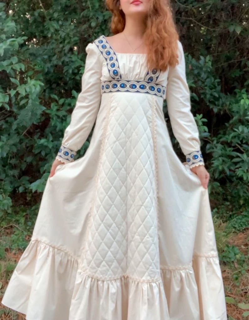 Puede incluir: Vestido largo color crema con mangas largas, cuello cuadrado y corpi&ntilde;o acolchado. El vestido presenta detalles bordados en azul y blanco en el escote, la cintura y los pu&ntilde;os. La falda tiene un dobladillo con volantes, creando una silueta fluida.