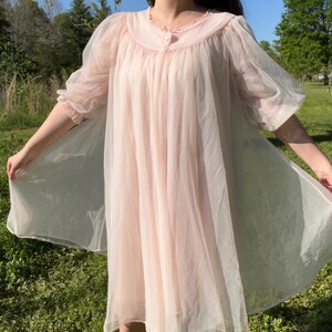 May include: A light pink vintage nightgown with a round collar and sheer, puffy sleeves. The nightgown has a flowing, A-line silhouette and is made of a lightweight, semi-transparent fabric. The collar is trimmed with delicate ruffles.