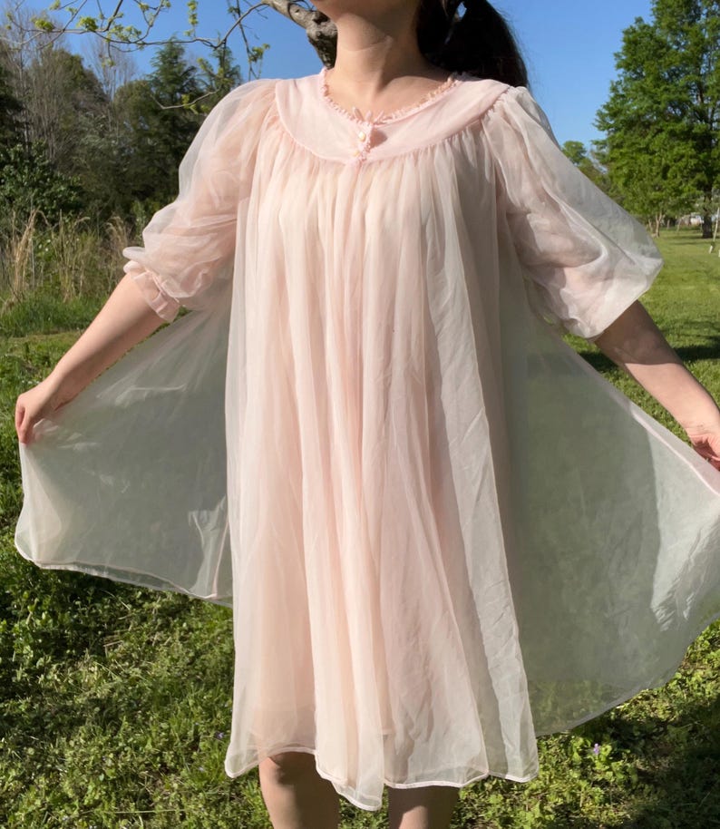 May include: A light pink vintage nightgown with a round collar and sheer, puffy sleeves. The nightgown has a flowing, A-line silhouette and is made of a lightweight, semi-transparent fabric. The collar is trimmed with delicate ruffles.