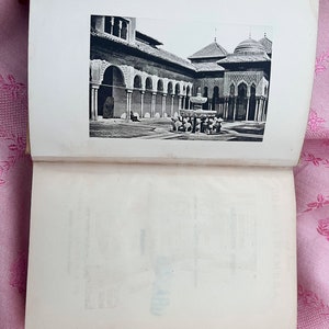 May include: An open book displaying a black and white photograph of an architectural courtyard. The courtyard features arched colonnades, a fountain, and domed structures. The image is printed on a white page.