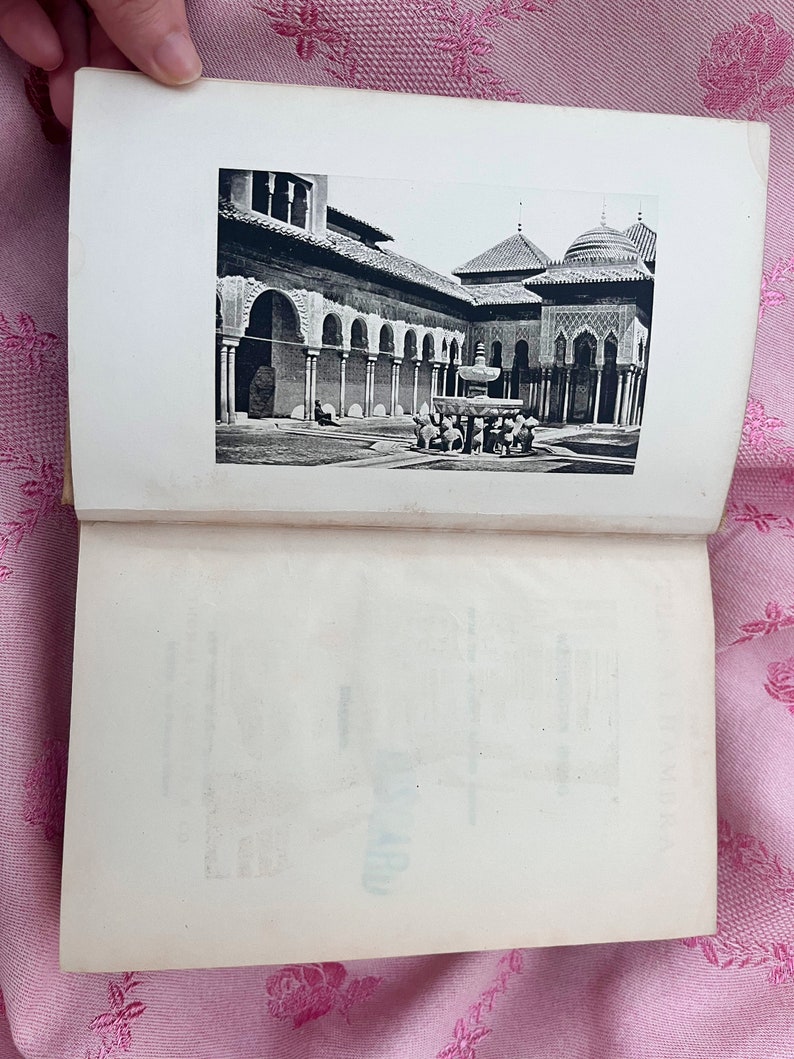 May include: An open book displaying a black and white photograph of an architectural courtyard. The courtyard features arched colonnades, a fountain, and domed structures. The image is printed on a white page.