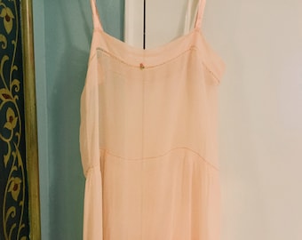 Puede incluir: Un camisón color melocotón con tirantes finos cuelga de una percha acolchada. El vestido presenta un delicado bordado a lo largo del escote y un pequeño detalle floral. El dobladillo inferior tiene volantes en capas.