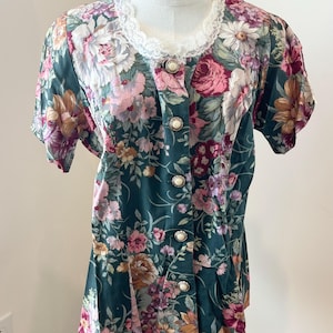 Puede incluir: Una blusa vintage con estampado floral en tonos verdes, rosas y blancos. La parte superior de manga corta presenta una parte delantera abotonada con botones nacarados y un delicado cuello de encaje. La blusa se muestra en un maniquí.