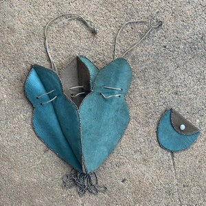May include: Handmade teal leather pouch and small coin purse. The pouch has a unique leaf-like design with silver stitching and a drawstring closure. The coin purse is teal and brown with a button closure. Both are on a textured surface.
