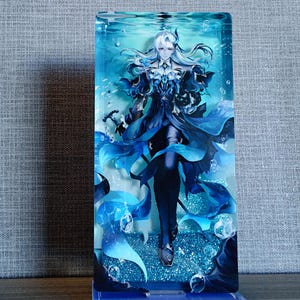 Peut inclure: Un présentoir rectangulaire en acrylique avec une illustration de personnage en nuances de bleu et de blanc, sur un fond aquatique avec des bulles. Le personnage a de longs cheveux blancs et une tenue bleue détaillée. La base du présentoir porte le texte "Genshin Impact".