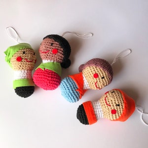 Puede incluir: Cuatro adornos de muñecas de ganchillo hechos a mano con caras sonrientes y varios atuendos. Las muñecas tienen cabello blanco, negro y marrón, y visten ropa verde, roja, naranja y azul. Cada muñeca tiene una cuerda blanca para colgar.
