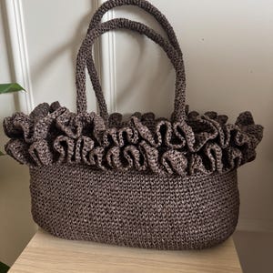 May include: A brown woven tote bag with a ruffled top edge and two matching handles. The bag is rectangular with rounded corners and sits on a light-colored wooden surface. The bag's texture appears to be tightly woven.