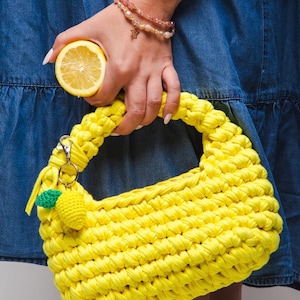 Może przedstawiać: Jasnożółta, ręcznie szydełkowana torebka z pasującą rączką. Torebka posiada mały cytrynowy urok i srebrny zapięcie. W pobliżu trzymana jest połówka cytryny. Torba wykonana jest z grubej, teksturowanej przędzy.