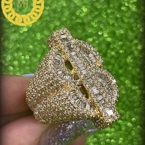 Könnte beinhalten: Ein goldener Ring mit einem großen, diamantbesetzten Dollarzeichen-Design. Der Ring ist mit Baguette-förmigen Diamanten besetzt.