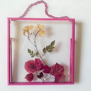 May include: A pink metal frame with a glass front, hanging from a chain, displays pressed flowers. The frame contains yellow and pink flowers.