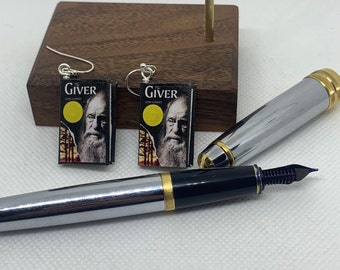 May include: A pair of earrings featuring miniature book covers of "The Giver" by Lois Lowry. The earrings are silver and the book covers are black with gold lettering and a yellow circle. The earrings are displayed on a wooden surface with a silver pen.