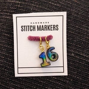 May include: Handmade stitch markers with the numbers 1 and 6 in a gradient of colors, attached to gold-colored clasps. The markers are displayed on a white card with the text "HANDMADE STITCH MARKERS". The background is a dark, textured fabric.