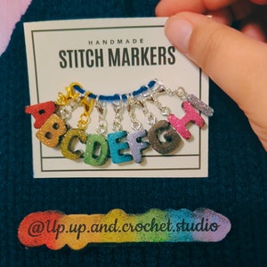 May include: Handmade stitch markers with glitter letters A through H. The letters are rainbow colored and have silver rings. The card reads "Handmade Stitch Markers" and "@Up.up.and.crochet.studio".