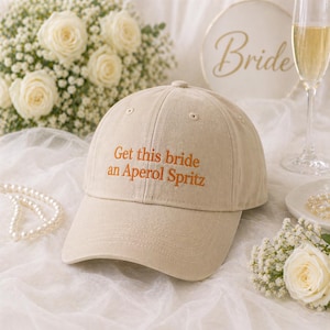 May include: A beige baseball cap with the embroidered text "Get this bride an Aperol Spritz" in orange. The cap is on a white surface with flowers, a pearl necklace, and a champagne glass in the background. A sign that says "Bride" is also in the background.