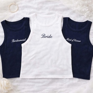May include: Three sleeveless tank tops featuring crew necklines. The centre white tank top has the word Bride embroidered in black. The two navy blue tank tops on either side feature the words Bridesmaid and Maid of Honour in white script.