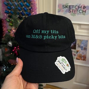 May include: Black baseball cap featuring teal embroidery with the words "Off my tits on M&S picky bits". A sticker on the brim reads "Love your order?". The cap is held by a hand.