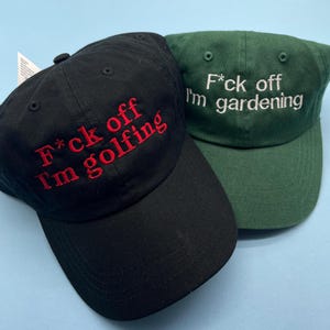 May include: Two baseball caps, one black and one green. The black cap has red embroidered text that reads "F*ck off I'm golfing." The green cap has white embroidered text that reads "F*ck off I'm gardening."
