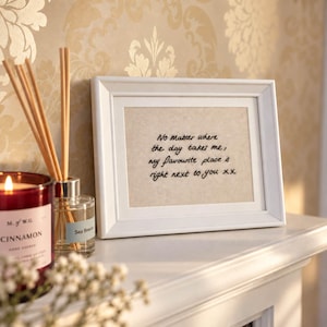 May include: A white picture frame containing a beige fabric insert with black embroidered text that reads: No Matter where the day takes me, my favourite place is right next to you xx. The frame is positioned next to a glass reed diffuser.
