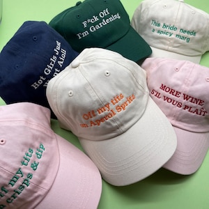 May include: Several baseball caps in shades of navy, green, and pink. Each cap has embroidered text, including phrases like "F*ck Off I'm Gardening," "More Wine S'il Vous Plaît," and "Off my tits on Aperol Spritz." A fun, casual accessory.