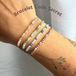 Puede incluir: Cuatro delicadas pulseras de cuentas en colores pastel: melocotón, verde, azul y malva. Las pulseras están ensartadas en una fina cadena dorada. El texto "Bracelet Code Secret" está escrito en cursiva marrón.
