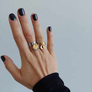 Peut inclure: Une main portant deux bagues en or avec des breloques et une bague en argent avec un breloque. Les bagues sont sur le majeur. Les ongles sont peints en noir.