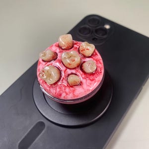 May include: A circular phone grip featuring a realistic pink gum-like base embedded with six off-white human-like teeth. The surface of the gums is textured with red, blood-like splatters and veins, creating a detailed, anatomical appearance.