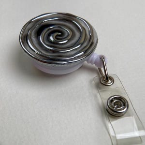 以下が含まれることがあります： グレー、黒、白の光沢のある盛り上がった渦巻き模様が特徴の円形バッジリール。リールは白いプラスチック製のハウジングに取り付けられており、透明なビニールストラップと銀色の金属製スナップボタン留め具が付いています。